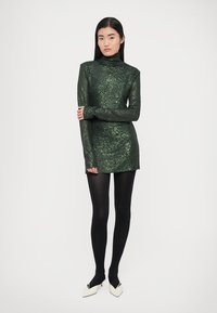 Zielona cekinowa mini sukienka z golfem i długimi rękawami, zestawiona z czarnymi rajstopami i białymi butami na obcasie.