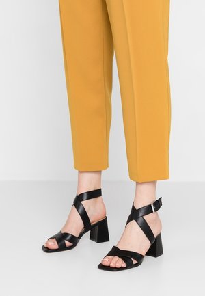 Czarne sandały na obcasie z paskami, z kwadratowym noskiem, wyposażone w kilka ukośnych pasków i regulowaną klamrę przy kostce, zestawione z musztardowymi spodniami.