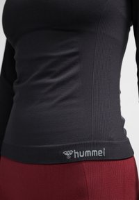 Close-up van een persoon die een aangesloten zwarte longsleeve draagt met het "hummel"-logo vlak bij de zoom en bordeauxrode geribbelde broek.