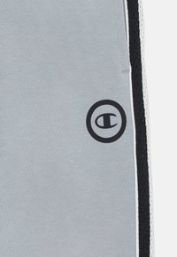 Pelēki sporta bikses ar melnu sānu paneļu, baltām akcentiem un melnu apļa logotipu uz augšstilba; gluda tekstūra un sportiska fit.