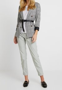 Žena v světle šedých kalhotách, bílém topu, šedém kostkovaném saku s páskem a černých mokasínech, stojící na jednobarevném bílém pozadí.