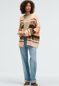 Soft, striped cardigan in pink, brown, and white, with a loose fit and long sleeves. Paired with light blue, slightly flared jeans.