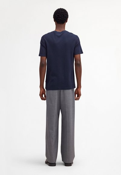 Mand stående med ryggen til, iført en navyblå kortærmet t-shirt, grå viddebukser og sorte sko mod en hvid baggrund.