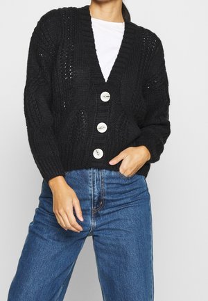 Osoba nosící černý pletený cardigan s velkými bílými knoflíky přes bílou košili, zapnutou do vysokých modrých džínů, ruce v kapse.