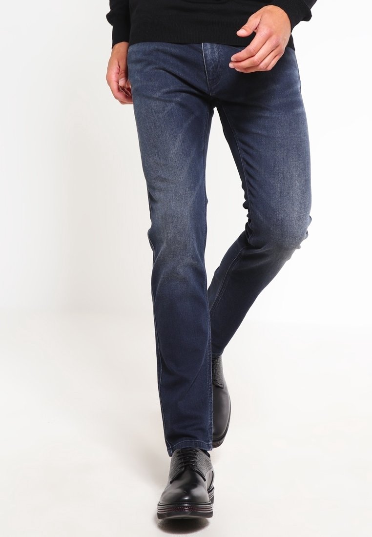 Vaqueros de mezclilla azul oscuro con un diseño ajustado. Textura suave, un ligero desvanecimiento y estilo de cinco bolsillos. Combinados con zapatos de vestir negros.