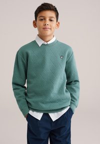 Zelená frotté mikina s textúrovaným šesťuholníkovým vzorom, rebrovanými manžetami a okrúhlym výstrihom, nosená cez bielu košeľu s golierom.