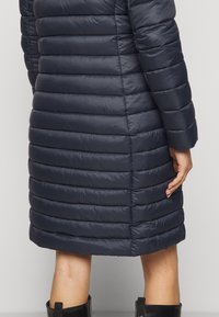Mørkeblå dunjakke med vandret quiltning, i mellemlang længde, med en glat tekstur og let glans. Bagdetaljen inkluderer en sideslids.