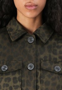 Leopardprint jasje in donker groen en bruine tinten, met een puntige kraag, twee borstzakken en vier zwarte knopen. Zachte stoffen textuur.