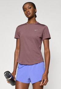 Μωβ αθλητικό σορτς και εφαρμοστό καφέ κοντομάνικο μπλουζάκι, με μικρό ασημένιο λογότυπο, κατασκευασμένα από ελαφρύ, διαπνέον ύφασμα.