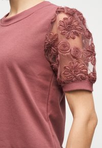 Μωβ τοπ με διάφανο φουσκωτό μανίκι που διαθέτει κεντημένα λουλουδάτα και στροβιλιστά σχέδια, ριμπ μανσέτα και στρογγυλή λαιμόκοψη, φορεμένο σε γυμνό χέρι.