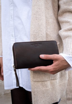 Brown leather wallet with a zip closure, featuring a textured surface and embossed brand name. Held in a hand, set against neutral clothing.
