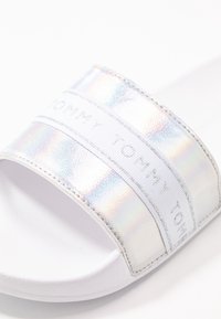 Infradito olografico con una fascia bianca testurizzata con branding in rilievo e una suola morbida bianca. Design standard con punta aperta.