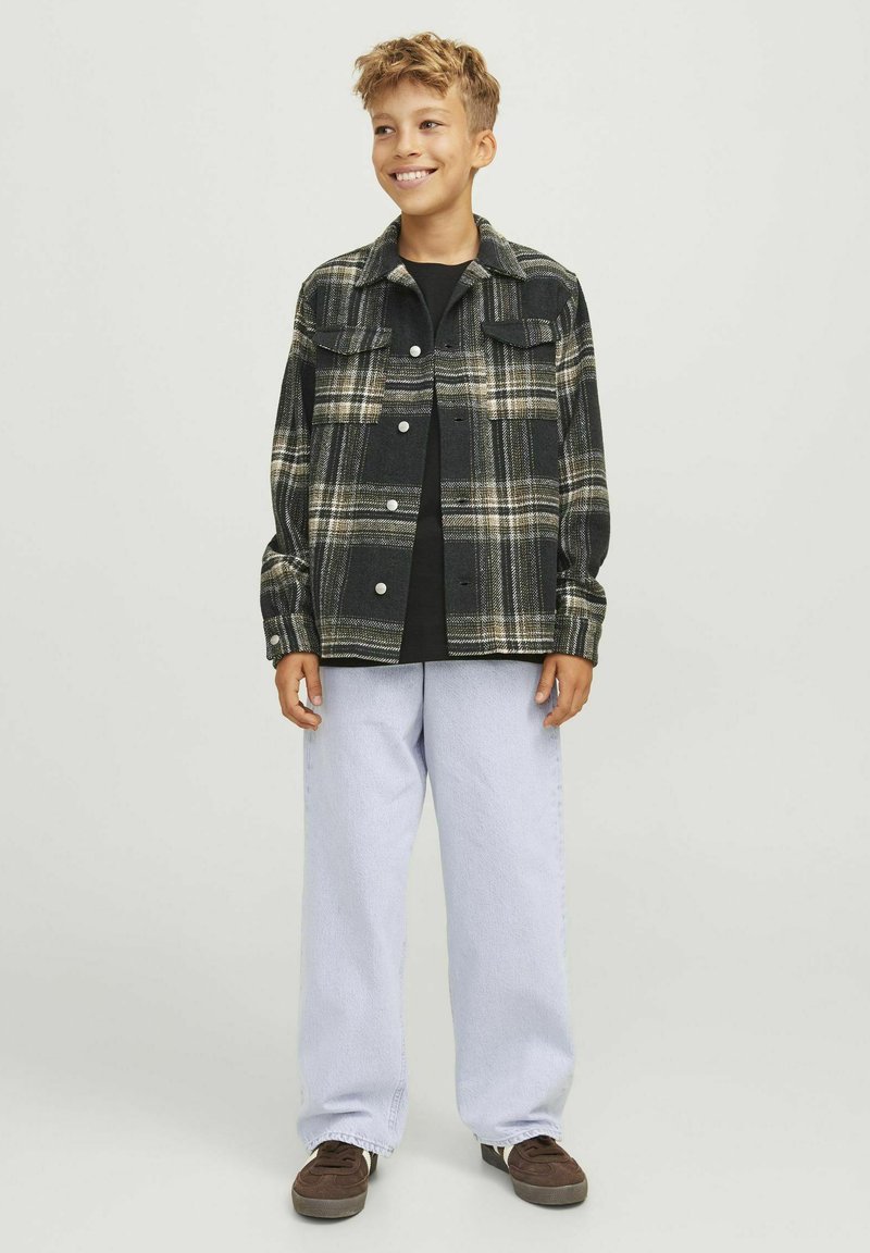 Camisa de franela a cuadros en negro y beige, con dos bolsillos en el pecho y cierre de botones. Combinada con pantalones holgados de color azul claro y zapatos marrones.