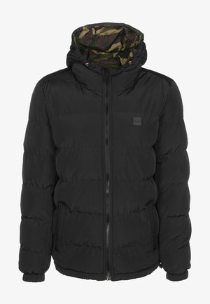 Henkilö pukeutuneena vihreään, ruskeaan ja mustaan maastokuvioiseen hupulliseen takkiin, jossa on joustavat hihansuut, mustaan paitaan ja tummanharmaisiin farkkuihin, nähtynä takaa.