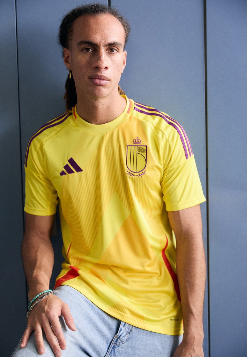 Yellow football jersey with red accents, diagonal stripes, and purple shoulder bands. Fabric appears lightweight, fitted, and sports a logo.