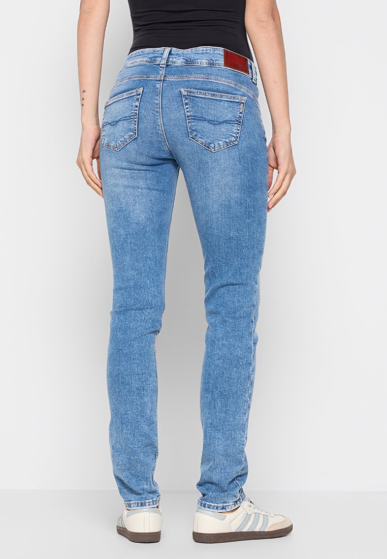 Schmale blaue Jeans mit einer ausgewaschenen Optik, zwei Gesäßtaschen und einem roten Lederpatch am Bund, kombiniert mit weißen Sneakers.