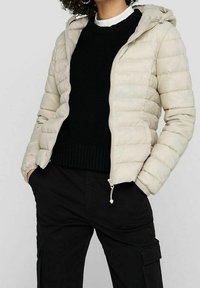 Doudoune beige avec capuche, équipée d'une fermeture éclair frontale, motif matelassé horizontal et texture légère. Portée sur un pull noir.