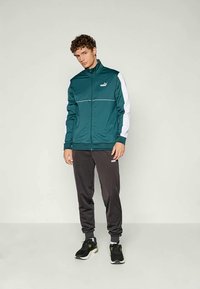 Mėlynas zip-up džemperis su baltais akcentais ir logo, derinamas su tamsiai pilkomis sportinėmis kelnėmis ir juodais sportiniais batais. Lygus audinio paviršius.