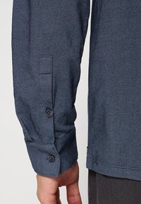 Overhemd met lange mouwen in donkerblauwe stof met een diagonale visgraatpatroon; heeft een getailleerde pasvorm en manchetten met knopen.