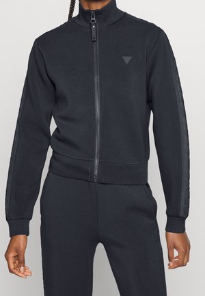 Persoon draagt een zwart zip-up trainingsjack met bijpassende broek, met een klein driehoekig logo op borst en been, en getextureerde strepen op de mouwen.