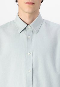 Chemise bleu clair à manches longues, à col pointu et texture lisse. Cinq boutons bordent la patte de boutonnage à l'avant, avec des détails de couture subtils.