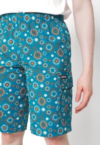 Niebieskie szorty z wzorami słońca i kwiatów, elastyczny pas i kieszeń boczna. Wykonane z lekkiego materiału. Metka marki na kieszeni.