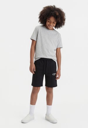 Šypsantis vaikas su garbanotais plaukais, dėvintis šviesiai pilką marškinėlį, juodus šortus su raišteliu, baltas kojines ir baltus slip-on batus, stovintis priešais baltą foną.