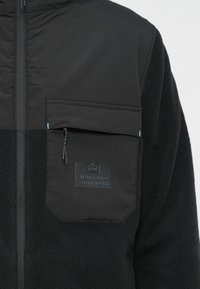 Sort fleecejakke med en glat frontpanel og teksturerede ærmer. Har en lynlåslomme på brystet og et logo-patch på lommen.