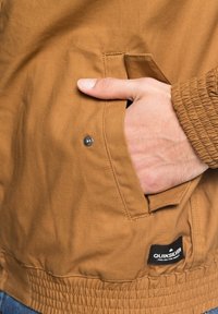 Hnědá bunda s текстuřeným povrchem, se služečnou kapsou, elastickým pasem a logem Quiksilver blízko lemu.