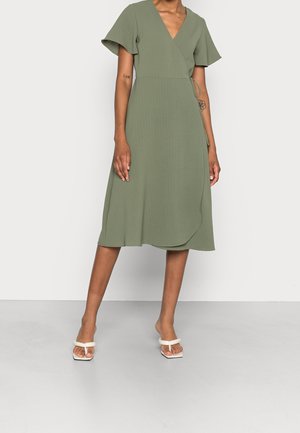 Femme portant une robe portefeuille vert olive à manches courtes et des sandales blanches à talons, debout devant un fond clair uni.