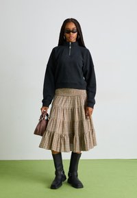 Sudadera negra con cremallera, falda de cuadros en tonos beige y marrón con volantes, botas altas negras hasta la rodilla, bolso pequeño marrón y gafas de sol. Fondo verde.