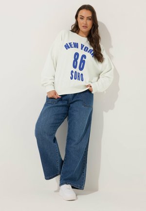 Weißer Kapuzenpullover mit blauer Aufschrift "NEW YORK 86 SOHO" und locker sitzenden blauen Jeans. Model trägt weiße Sneakers und steht vor einem schlichten Hintergrund.