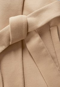 Beige kabát textúrált, puha anyagból. Kiemelkedő megkötő díszítéssel és letisztult vonalakkal rendelkezik. Minimalista dizájn, diszkrét zsebekkel.