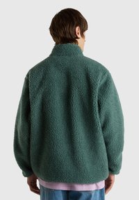 Fleecejacka i mörkgrön med en texturerad, fluffig yta. Har hög krage och lös passform, bärs över en ljusrosa skjorta och jeans.