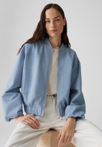 Veste aviateur bleue avec un zip à l'avant, un col côtelé et des manches volumineuses, associée à un haut en tricot léger et un pantalon de couleur crème.