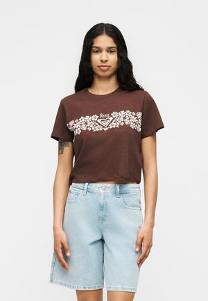 Femme aux longs cheveux foncés portant un t-shirt court marron avec un imprimé floral blanc et un short en jean bleu clair, debout devant un fond uni.