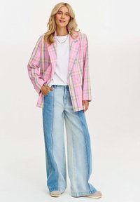Ružový kockovaný dvojradový blejzer s modrými akcentmi, v kombinácii so širokými denimovými džínmi vo svetlej a stredne tmavej farbe a béžovými topánkami.