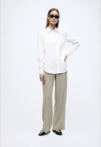 Λευκό πουκάμισο με κουμπιά, μπεζ παντελόνι wide-leg, μαύρα παντόφλες. Το μοντέλο φοράει μαύρα γυαλιά ηλίου και στέκεται μπροστά σε ένα απλό φόντο.