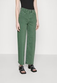 Vaqueros amplios verdes de denim, con bolsillos frontales, costuras visibles y una cintura alta, combinados con sandalias de tacón cuadrado negras.