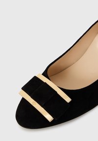 Pump in suede nero con punta affusolata, doppi accenti orizzontali dorati e dettaglio a nastro nella parte anteriore. Interno liscio con leggera texture.