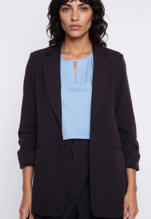 Kobieta w czarnej marynarce z podwiniętymi rękawami, na jasnoniebieskiej bluzce z przednim rozcięciem, stoi na jednolitym tle.