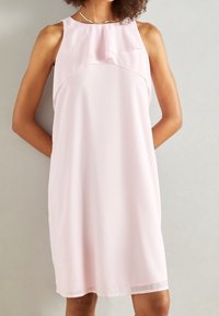 Donna che indossa un vestito senza maniche di un leggero rosa chiaro, lungo fino al ginocchio, con dettagli a strati e volant sul petto, in piedi davanti a uno sfondo neutro.