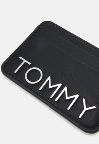 Černý kapsička na karty vyrobená z texturované kůže. Obsahuje stříbrné písmo "TOMMY" a sloty na karty po horním okraji.