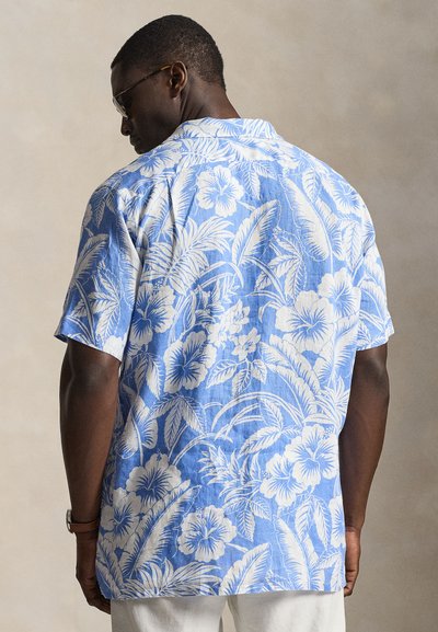 Moški, obrnjen stran, oblečen v modro srajco s kratkimi rokavi z belo tropsko cvetlično vzorcem in svetle hlače, na nevtralni ozadju.