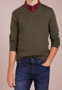 Man i olivgrön V-halsad tröja över en vinröd skjorta, med ena handen i fickan på sina mörkblå jeans.