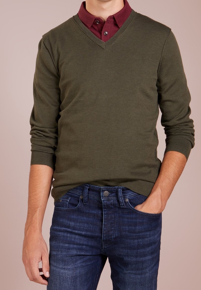 Man i olivgrön V-halsad tröja över en vinröd skjorta, med ena handen i fickan på sina mörkblå jeans.