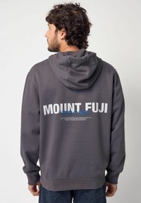 Sweatshirt à capuche gris avec "MONT FUJI" en blanc dans le dos, des détails en texte bleu et un tissu doux et texturé.