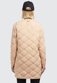 Chaqueta acolchada de color beige claro con una textura suave, que presenta un patrón de ondas y mangas largas, vista desde la parte posterior, combinada con una gorra negra.
