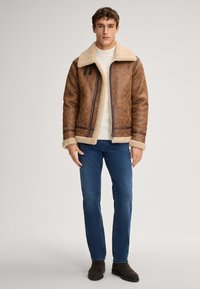 Veste en cuir synthétique marron avec col et poignets en peau de mouton, fermeture éclair et deux poches latérales, associée à un jean bleu et des bottes noires.