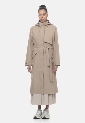 Donna con trench coat beige con cintura e cappuccio sopra una gonna plissettata e sneakers bianche, in piedi davanti a uno sfondo chiaro e uniforme.